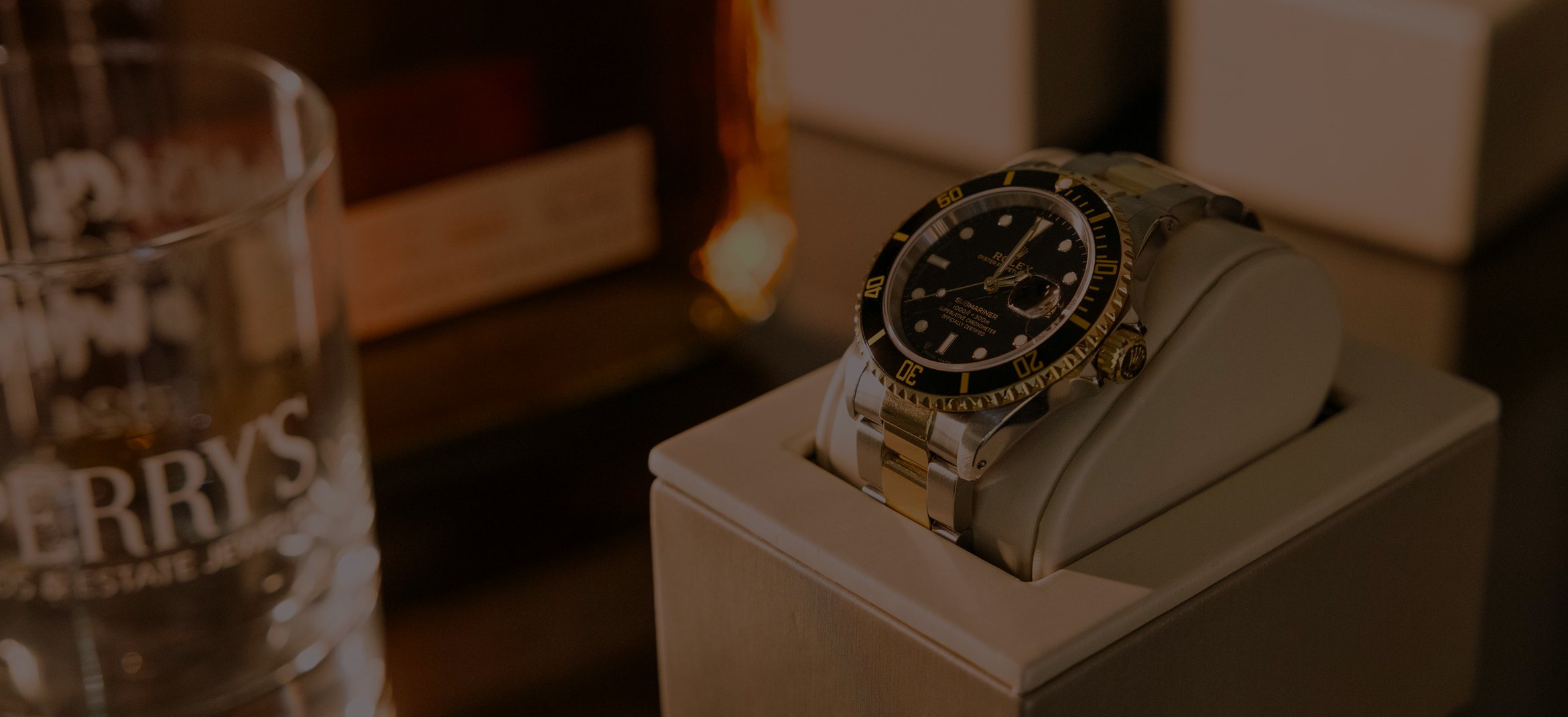 A two-tone Rolex Submariner on a display cushion beside an etched Perry’s whiskey glass