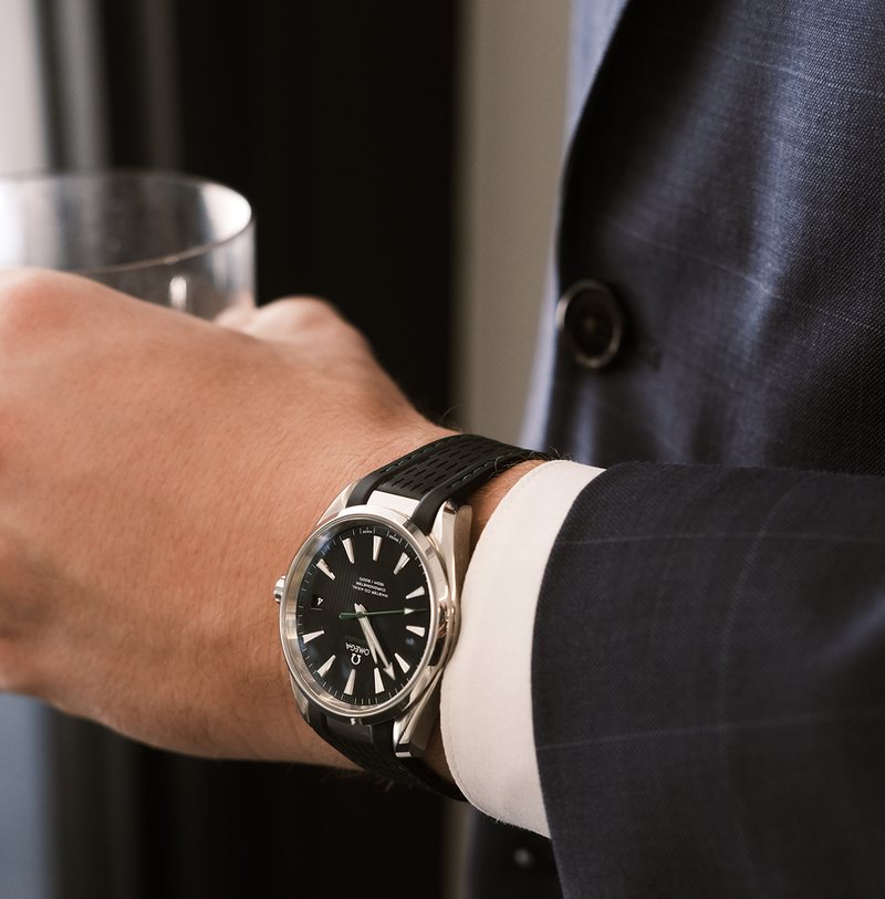 A guest in a navy suit holding a whiskey glass, wearing a black-dial steel watch